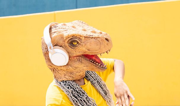 mulher com máscara t-rex, e fones de ouvido escuta música em uma parede amarela e azul - Foto, Imagem