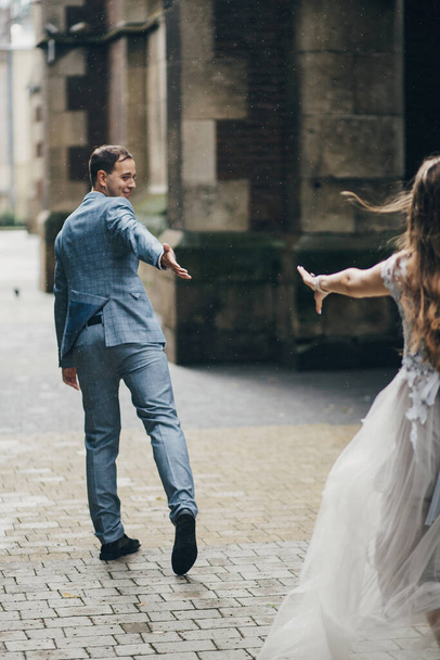 Stylish bride and groom running on background of old church in rainy street. Provence wedding. Beautiful emotional wedding couple having fun in rain in european city. - Фото, изображение