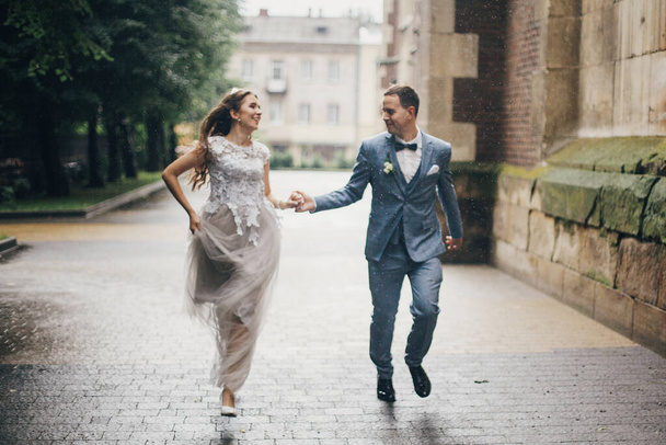 Verschwommenes Bild eines schönen emotionalen Hochzeitspaares, das im Regen in einer europäischen Stadt lächelt. Hochzeit in der Provence. Schicke glückliche Braut und Bräutigam laufen auf dem Hintergrund der alten Kirche in der verregneten Straße. - Foto, Bild