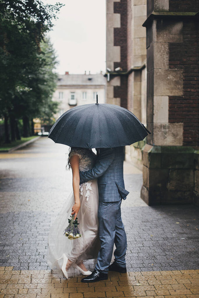 Stilvolles Brautpaar küsst sich unter einem Regenschirm vor dem Hintergrund der alten Kirche im Regen. Hochzeit in der Provence. Schönes Hochzeitspaar, das sich unter einem schwarzen Regenschirm in der verregneten Straße umarmt. Romantischer Moment - Foto, Bild