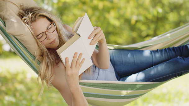 Lächelnde blonde Frau mit Brille liest ein Buch, entspannt auf der Hängematte im Garten, Freizeit- und Sommerferienkonzept - Foto, Bild