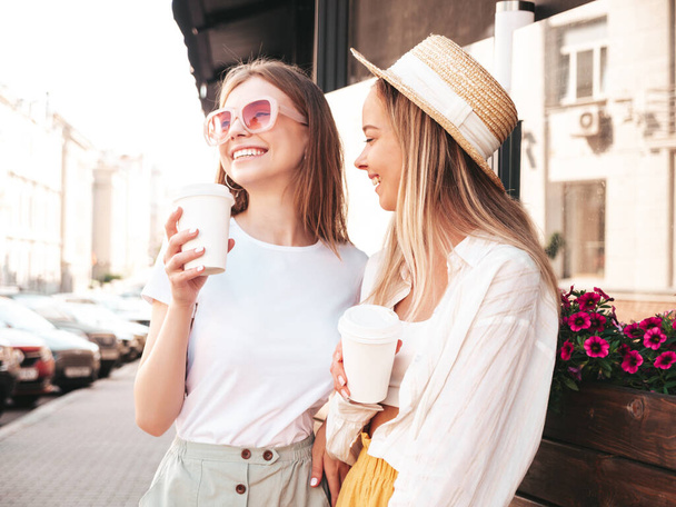 Two young beautiful smiling hipster female in trendy summer clothes.Sexy carefree women posing in the street. Positive pure models having fun at sunset. They drinking coffee or tea in plastic cup - Foto, Bild