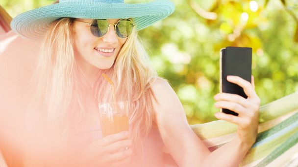 Smiling blonde woman with sunglasses using smartphone, relaxing on the hammock in garden, drinks a juice, free time and summer holiday concept for surf internet or chat with friends using social media - Photo, Image