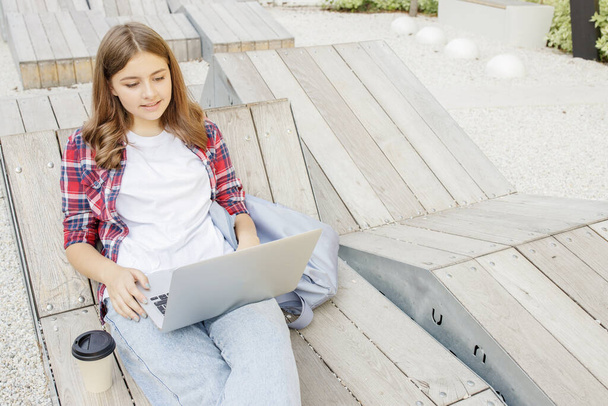 Joyeux adolescent, blogueur avec ordinateur portable sur le banc du campus. L'écolière est assise dans le parc à étudier en ligne. Mode de vie. Concept Wi-Fi dans les lieux publics, école moderne, enseignement à distance, achats en ligne. - Photo, image