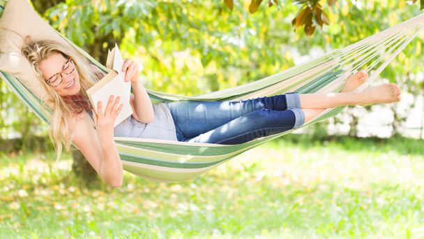 Lachende blonde vrouw met bril die een boek leest, ontspannen op de hangmat in de tuin, vrije tijd en zomervakantie concept - Foto, afbeelding