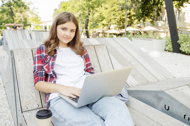 Menina adolescente feliz, blogueira com laptop no banco do campus. Escola menina senta-se no parque estudando online. Estilo de vida. Conceito de Wi-Fi em lugares públicos, escola moderna, educação a distância, compras on-line. - Foto, Imagem