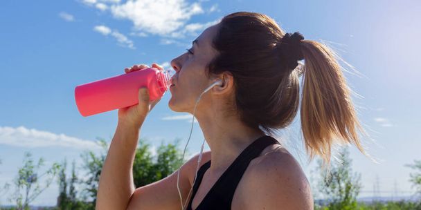 Young woman drinking water after running listening to music with headphones. Healthy lifestyle. Selective focus. - Photo, Image