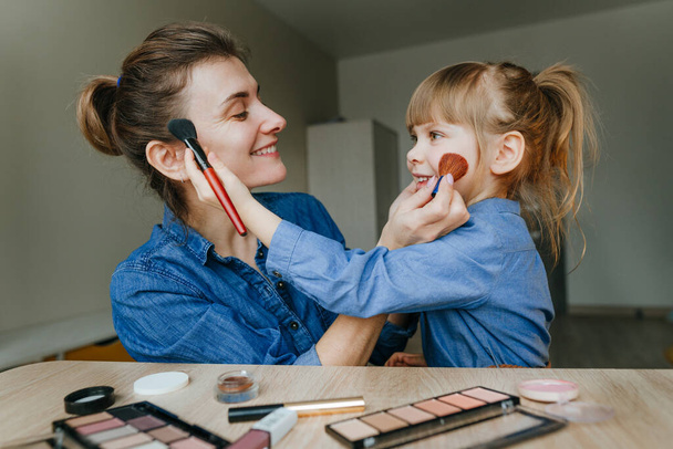 Portrait of young mother and her little daughter making make up each other with makeup brushes indoor. Family fun and beauty care concept. Selective focus. - Foto, Bild