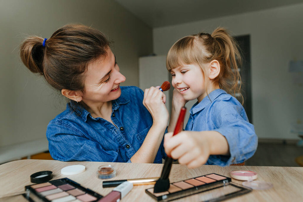 Portrait of young mother and her little daughter making make up each other with makeup brushes indoor. Family fun and beauty care concept. Selective focus. - Photo, Image