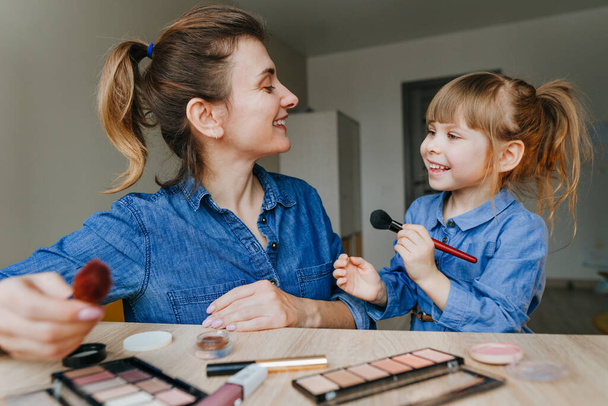 Portret van de jonge moeder en haar dochtertje die samen make-up maken met make-up borstels binnen. Familie plezier en schoonheidsverzorging concept. Selectieve focus. - Foto, afbeelding