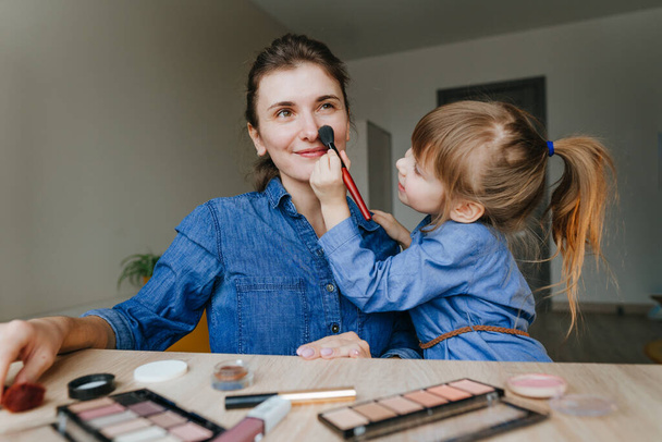 Portret van de jonge moeder en haar dochtertje die samen make-up maken met make-up borstels binnen. Familie plezier en schoonheidsverzorging concept. Selectieve focus. - Foto, afbeelding