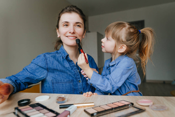 Kapalı alanda makyaj fırçalarıyla birbirlerini tamamlayan genç anne ve küçük kızının portresi. Aile eğlencesi ve güzellik konsepti. Seçici odak. - Fotoğraf, Görsel