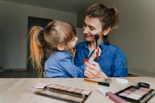 Porträt einer jungen Mutter und ihrer kleinen Tochter, die sich mit Schminkpinseln gegenseitig schminken. Familienspaß und Schönheitspflege. Selektiver Fokus. - Foto, Bild