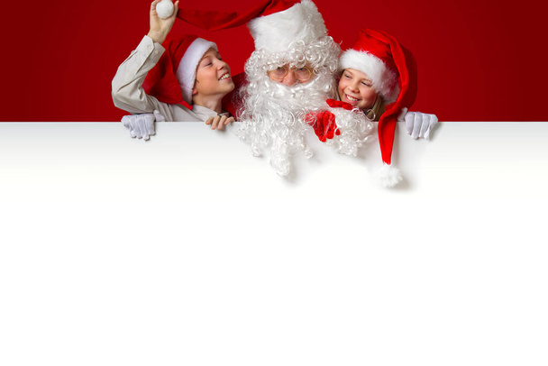 satna claus z zabawnymi dziećmi w czerwonych świątecznych kapeluszach gnome wyjrzał zza pustego białego transparentu - Zdjęcie, obraz