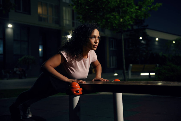Young mixed race woman athlete performing push-ups from a wooden bench on the sports ground and pumping chest and shoulder muscles during evening workout - Foto, imagen
