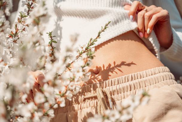 Μερική άποψη της όμορφης λεπτής γυναίκας σε ντελικάτα ρούχα ποζάρουν με τα χέρια κοντά στην κοιλιά της και με λευκά λουλούδια ανθισμένα λεπτή φωτογραφία, παστέλ χρώματα. - Φωτογραφία, εικόνα