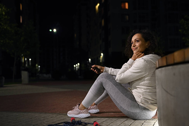 Lado retrato de comprimento total da bela mulher hispânica desfrutando de relaxamento após o treino à noite sentado no chão com telefone celular na mão e olhando para o lado - Foto, Imagem