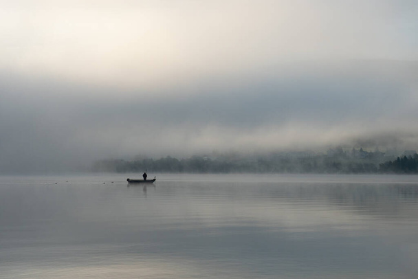 Una madrugada en Lac de Joux, Suiza: Un pescador solitario en un barco, la niebla todavía oculta el sol naciente - Foto, Imagen