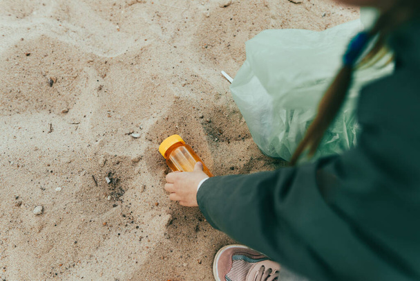 Крупный план ребенка, собирающего пластиковый мусор на песчаном пляже - бутылки, коробки и соломинки. - Фото, изображение