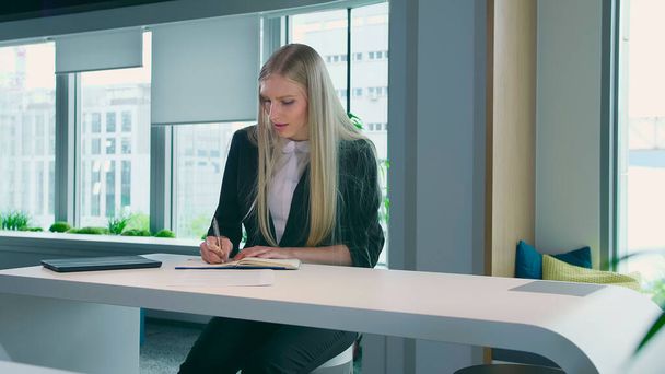 Элегантная женщина работает в стильном офисе. Современная блондинка в модном костюме сидит за столом в светлом современном офисе и пишет на газетах
. - Фото, изображение