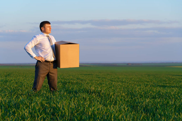 un hombre de negocios sostiene una caja de cartón y posa en un campo de hierba verde - concepto de negocio - Foto, Imagen