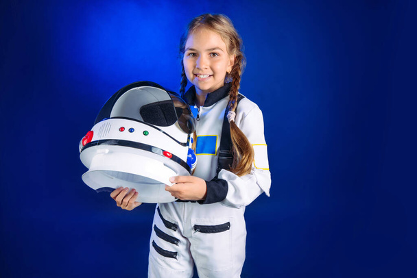 宇宙飛行士の衣装を着た10代の少女と宇宙服を着た暗い青の背景に立つヘルメット。宇宙服のかわいい女の子の肖像画. - 写真・画像