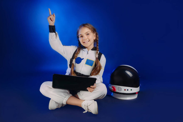 Teenager girl wearing astronaut costume using laptop sitting on dark blue background with copy space. Cute girl in spacesuit pointing up. - Photo, image
