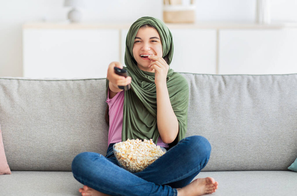 Unterhaltung zu Hause. Volle Länge eines muslimischen Teenagermädchens im Hijab mit Fernbedienung, das Popcorn isst und fernsieht - Foto, Bild