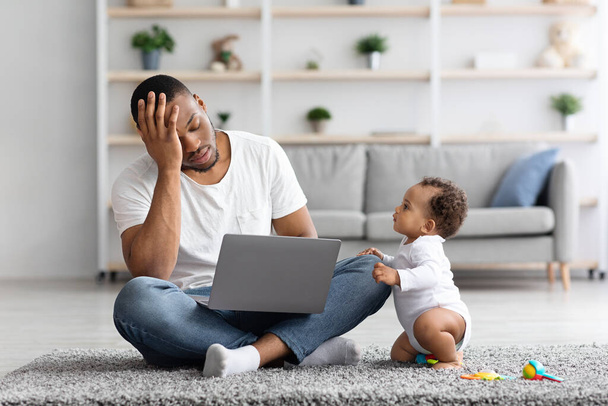 赤ん坊が彼を引き付ける間ノートパソコンと仕事をしようとしているストレス黒人男性 - 写真・画像