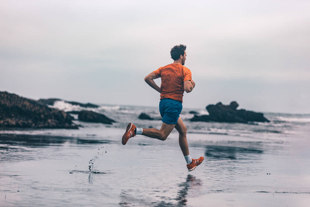 Exercise outdoor athlete man running on wet sand at beach training cardio sprinting fast. Profile of runner in sportswear clothes jogging - Fotoğraf, Görsel