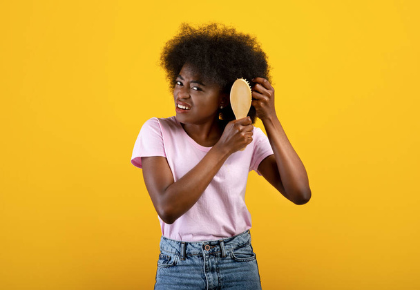 Concepto de peluquería doméstica. Mujer afroamericana frustrada tratando de cepillarse el cabello enredado, fondo amarillo - Foto, Imagen