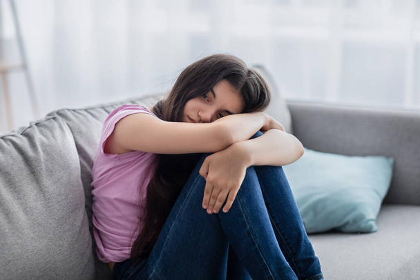 Δυστυχισμένο έφηβο κορίτσι που αισθάνεται κατάθλιψη ή απελπισία, κάθεται στον καναπέ στο σπίτι, πάσχει από πρόβλημα ή εφηβική κατάθλιψη - Φωτογραφία, εικόνα