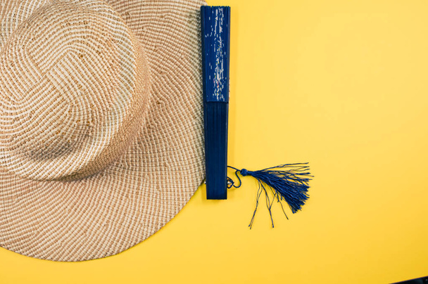 A round straw hat and a blue hand fan isolated on a yellow background with free space for text- concept for hot summer weather - Fotó, kép