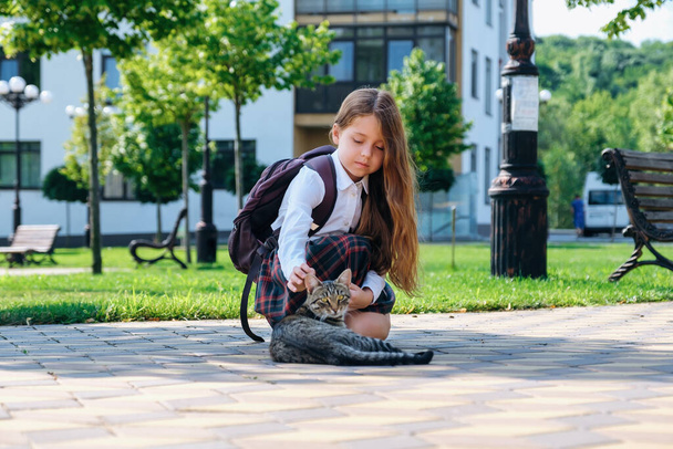 Roztomilá holčička v uniformě a s batohem hladí toulavou kočku na stree - Fotografie, Obrázek