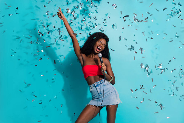 Photo of funky happy dark skin positive young woman sing song mic fall confetti isolated on blue color background - Photo, Image