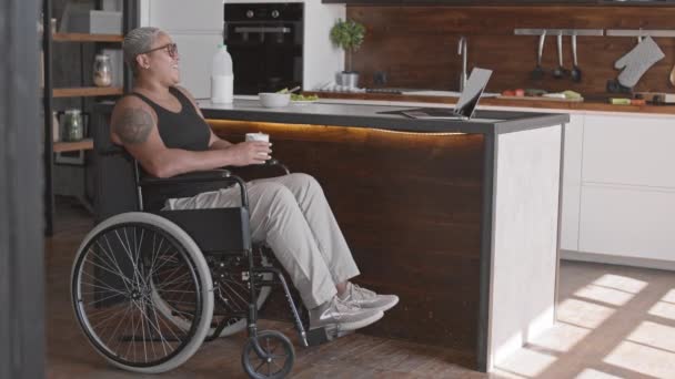 Boční pohled dlouhý mladý Smíšené závod žena sedí na invalidním vozíku, pití kávy z hrnku, sledování zábavné show na notebooku v kuchyni - Záběry, video