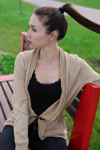 привлекательная брюнетка, сидящая на скамейке в парке. модная одежда, стильный вид - Фото, изображение