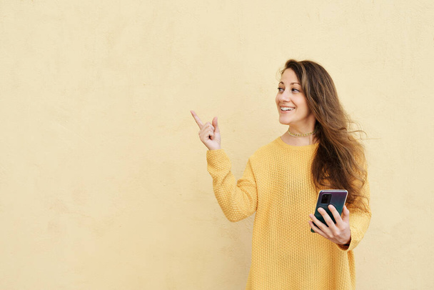 femme souriante pointant vers copier l'espace avec un smartphone à la main sur un fond jaune - Photo, image