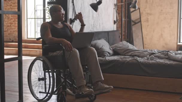 Volledig shot van kortharige vrouw zitten in een rolstoel in de slaapkamer, met laptop computer op de knieën, praten op luidspreker telefoon - Video