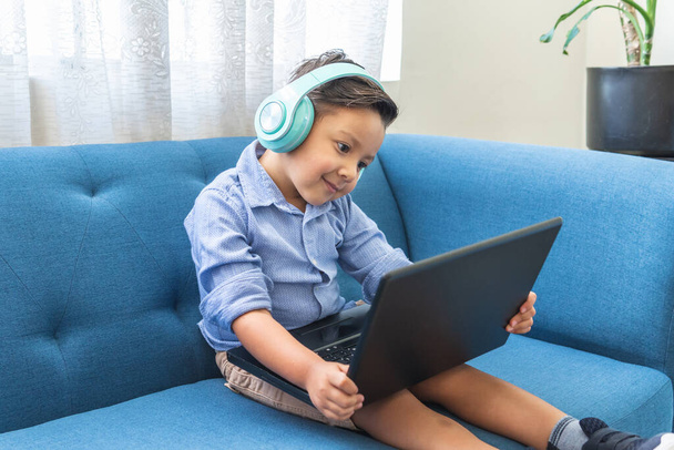 latynoski chłopiec ze słuchawkami w laptopie wideokonferencja siedzi na niebieskiej kanapie - Zdjęcie, obraz