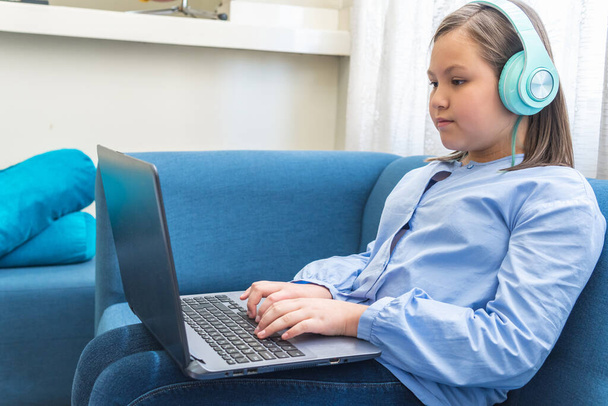 Menina loira com aparelhos auditivos usando seu laptop sentado em um sofá azul - Foto, Imagem