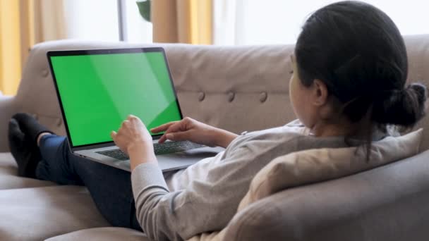 Green Mock-up ekranlı dizüstü bilgisayarda çalışan genç bir kadın. Onun rahat oturma odasındaki bir koltukta oturuyor. Omzunun üstünden. - Video, Çekim