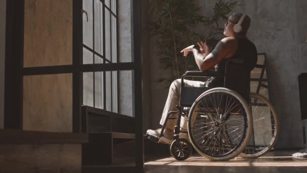 Full side view of young Mixed-Race woman wear over-ear ακουστικά κάθεται σε αναπηρική καρέκλα στο σπίτι, ακούγοντας μουσική και κινείται κατά μήκος - Πλάνα, βίντεο