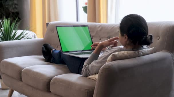 Young Woman at Home Works on a Laptop Computer with Green Mock-up Screen. Está sentada en un sofá en su acogedor salón. Sobre el hombro disparado - Imágenes, Vídeo