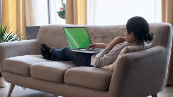 Mujer joven en casa acostada en un sofá funciona en una computadora portátil con pantalla verde simulada. Chica Usando Computadora, Navegando por Internet, Ver Contenido, Chatear en Redes Sociales con Amigos. - Imágenes, Vídeo