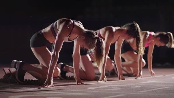 A row of runners womens crouch in the starting position before beginning to race. Females start with running shoes on the stadium from the start line in the dark with spotlights in slow motion. - Materiał filmowy, wideo