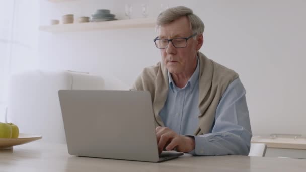 Seriöser Senior tippt auf Laptop, nimmt Brille ab und blickt irritiert auf Computer - Filmmaterial, Video