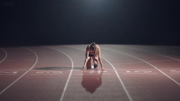 The woman at the start of the race gets into the pads, rises and runs in slow motion in the evening at the stadium. Female runner crouch in the starting position before beginning to race. - Séquence, vidéo