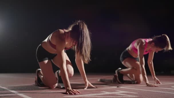 A row of runners womens crouch in the starting position before beginning to race. Females start with running shoes on the stadium from the start line in the dark with spotlights in slow motion. - Filmagem, Vídeo