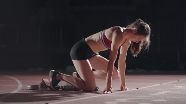 The woman at the start of the race gets into the pads, rises and runs in slow motion in the evening at the stadium. Female runner crouch in the starting position before beginning to race. - Imágenes, Vídeo